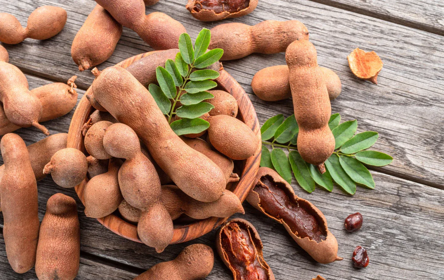 Fresh tamarind pods in a wooden bowl with green leaves on a rustic wooden table.