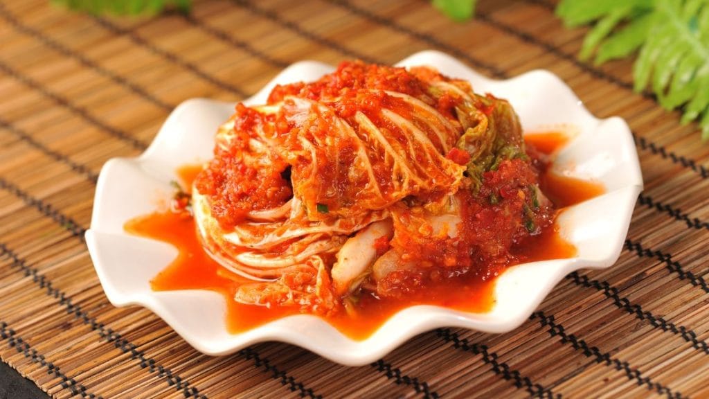 A white plate of kimchi, a Korean fermented vegetable dish with spicy red sauce, on a bamboo mat.